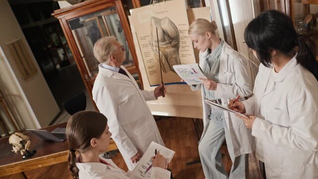 Senior Caucasian male professor explaining knee anatomy to diverse young adult female medical students taking notes in classroom. High angle shot ideal for medical education and university promos