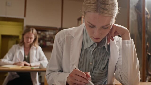 Young adult Caucasian female medical student wearing lab coat writing in notebook during lecture and looking up. Perfect for medical education and university recruitment campaigns