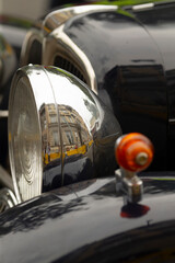 Close-up view of a polished chrome headlamp and glossy black fender of a vintage car. The curved metal surface reflects an urban street scene with buildings © Viktar