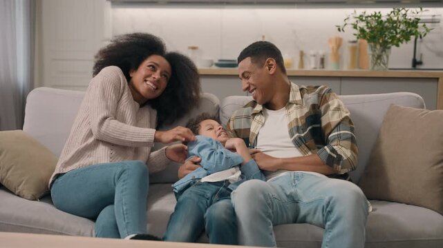 Playful African American family parents tickling son on bed in living room joyful mother mom father dad male man hug sincere laughing funny game ticklish having fun little boy child kid cuddle indoor