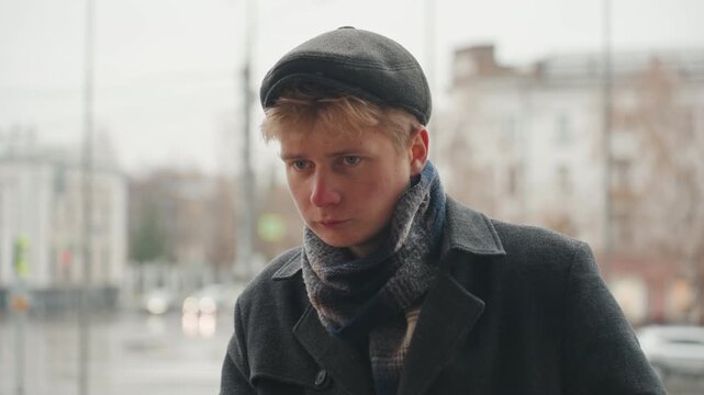 Introspective white creator on rainy street, closeup portrait showing contemplative young white male writer creator wearing flat cap and scarf, soft overcast light, blurred city background, muted