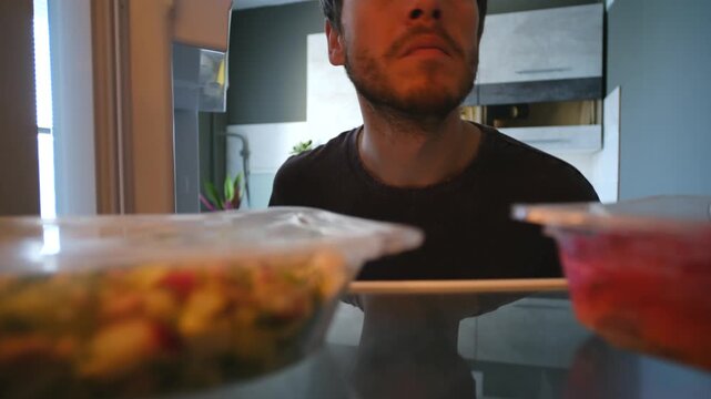 Hungry man opening fridge looking for food, seeing only pre packaged meals in plastic containers, closes fridge disappointed taking nothing. Hungry man closing fridge empty handed after searching food
