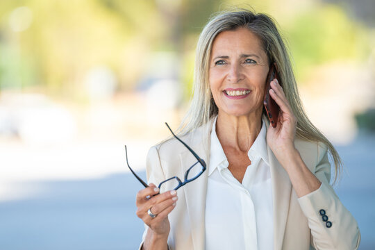 Smiling mature Caucasian businesswoman talking on smartphone outdoors, confident and cheerful expression, natural sunlight, blurred background, professional lifestyle portrait