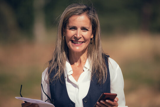 Smiling confident Caucasian businesswoman with long brown hair standing outdoors, holding papers and glasses, looking at camera with genuine happy expression in natural sunlight
