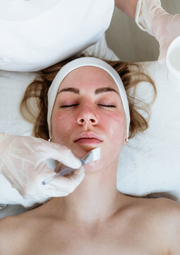 Cosmetologist applying exfoliating mask with brush on client's face during rejuvenating skincare procedure in wellness salon.