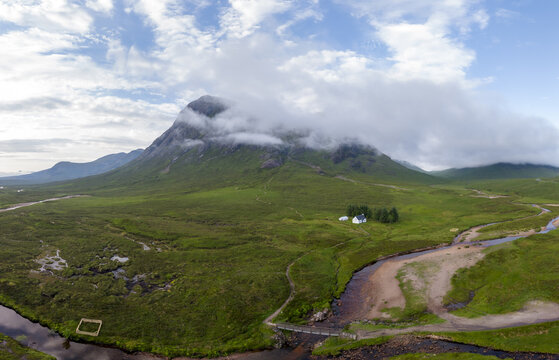 Aerial view of Buchaille Etive Mor a mountain in Glen Coe partially shrouded in clouds, lush green hills, a winding river, and a small white house in Scotland.