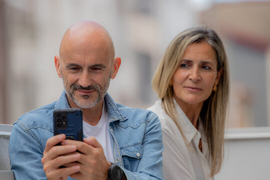 caucasian couple sitting outdoors, man smiling at smartphone while woman watches with suspicious expression, relationship tension concept