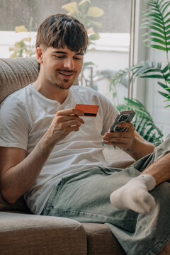Smiling young man sitting on sofa at home, holding credit card and looking at smartphone screen, making online payment or shopping on mobile phone in living room