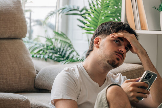 Stressed young Caucasian man holding smartphone, looking up with worried, frustrated expression while sitting indoors. Concept of stress and problems with technology or communication
