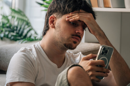 Stressed young man sitting on sofa looking at smartphone with worried expression. Anxious male reading mobile phone at home, feeling overwhelmed and stressed