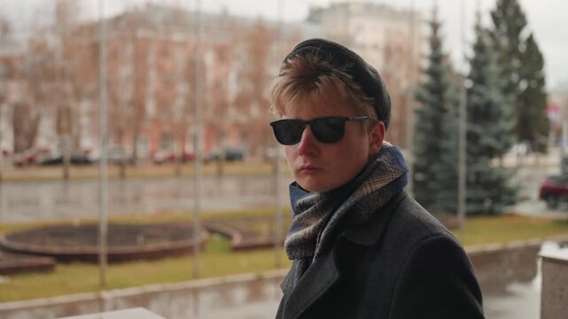 White creator in rainy plaza, contemplative portrait of white male creator wearing beret, sunglasses, scarf and coat, looking across fountain and wet pavement, moody cinematic tones, shallow depth