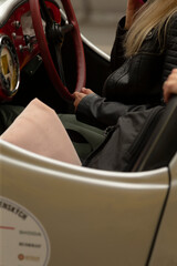 Obraz premium Partial view of a woman sitting in the driver’s seat of a vintage convertible. Visible are her crossed legs, black quilted leather jacket, hands on the