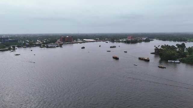 Drone view of houseboats moored along the shore of Punnamada Lake in Alappuzha, Kerala, set against green landscapes and buildings under a cloudy sky.