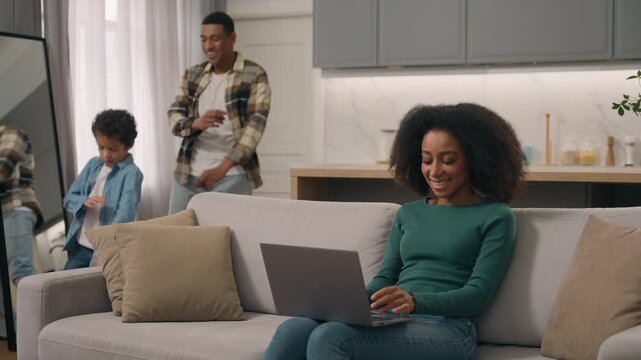 Successful African American woman mom mother freelancer using laptop notebook online work while father and little boy child son kid dancing music indoors apartment happy family business technology