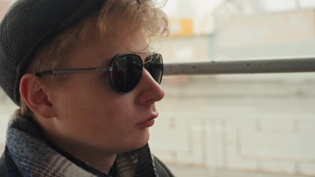 Young white creator adjusts sunglasses on cold urban platform, dons flat cap and dark round lenses while gripping metal rail and fixing steady gaze frames suggest director, model, and traveler