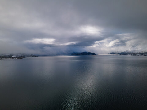 Aerial view of Malangen fjord with snow-covered mountains and dramatic cloudy skies over the dark water in Troms, Norway.