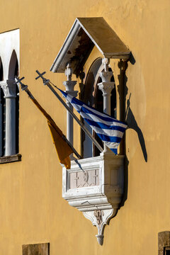 Flags hang from a decorative balcony on a yellow wall in Rhodes during bright daylight hours