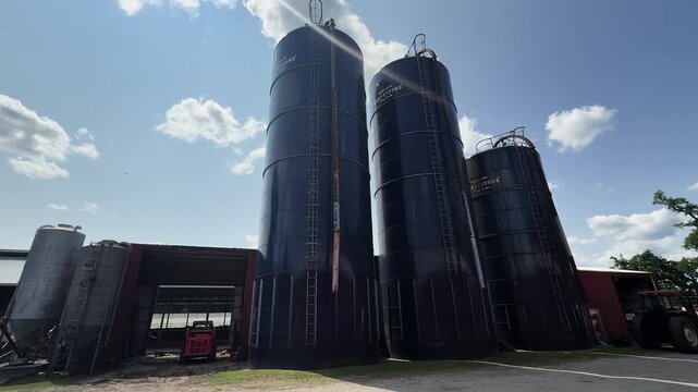 Tall grain silos stand beside farm buildings under a bright blue sky with clouds