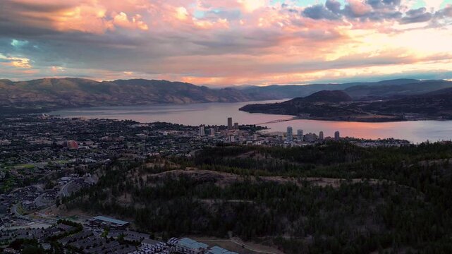 Cinematic aerial shot of Kelowna city panorama at sunset, showing Knox Mountain in foreground, Okanagan Lake, and the bridge under a dramatic red dusk sky.