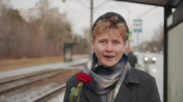 Young white man at tram stop hopeful poet creator offering red rose, knit cap and scarf, wool coat, shy smile, overcast urban background, cinematic closeup, candid street storytelling, heartfelt mood.
