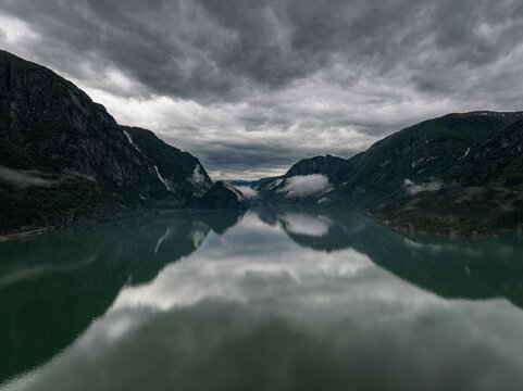 Aerial view of the calm waters of a fjord reflecting dramatic dark clouds and steep mountains in Odda, Vestland, Norway.
