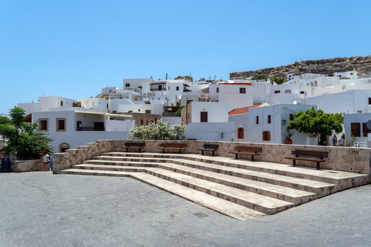 White buildings and steps in Lindos town square with benches and trees during the day