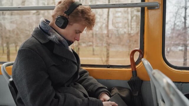 Young white creator on bus commute, wearing headphones and winter coat, scrolling smartphone, gazing through window at park, soft natural light, quiet reflective mood, candid documentary vibe, hands