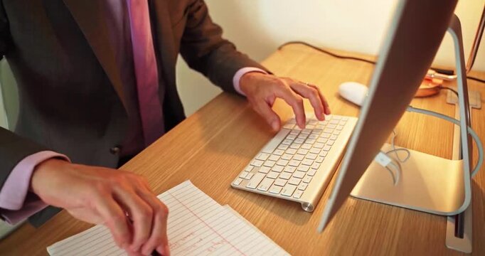 Businessman in brown suit typing on white keyboard then writing notes on lined paper pad.