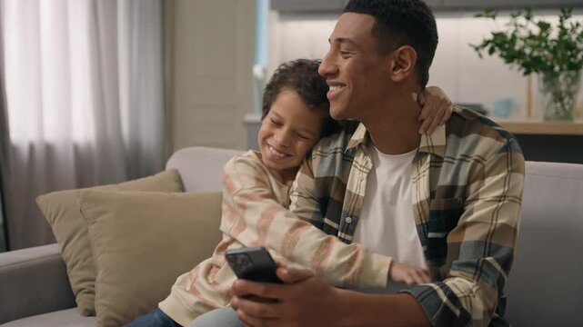 Joyful African American father playing online mobile phone game with little boy son child at home happy kid embracing daddy smartphone addiction work distraction sofa living room family happiness