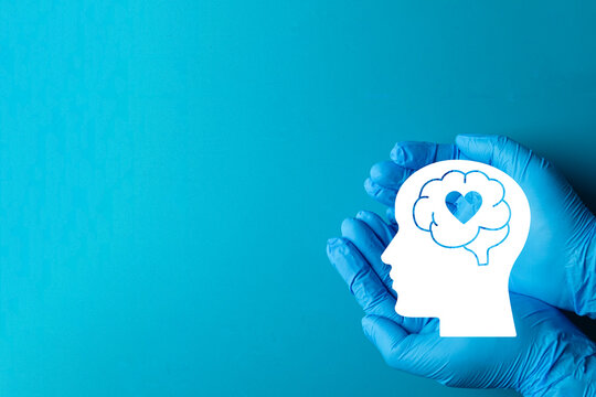 Mental health care and psychology concept. Doctor hands in blue medical gloves holding a human head silhouette with brain and heart on a solid blue background with copy space.