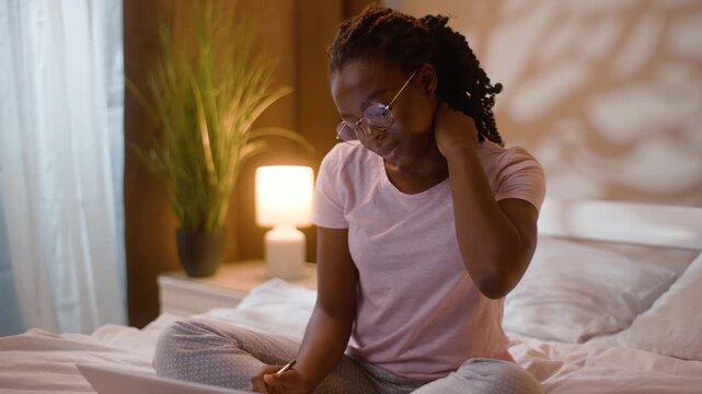 Tired African American woman with glasses working laptop on bed at home late night student neck pain rubbing ache doing homework typing writing notes female girl studying learning under lamp bedroom
