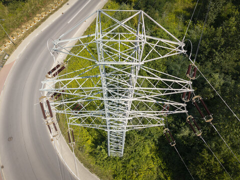 Aerial view of a high-voltage electricity pylon standing near a curved asphalt road and lush green forest in Poland.