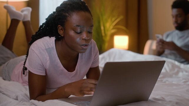 African American woman browsing internet typing laptop in bed boyfriend chatting mobile phone relaxed couple ethnic family weekend online computer work gadget addiction indoor home bedroom evening