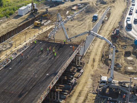 Aerial view of a bridge construction site with workers installing steel reinforcement bars on a concrete deck, assisted by a concrete pump truck, near a busy road in Poland.