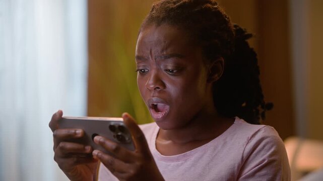 Shocked African American woman playing online game on smartphone at home bedroom night upset female gamer losing bet scared surprised user player frustrated girl mobile phone failure negative reaction