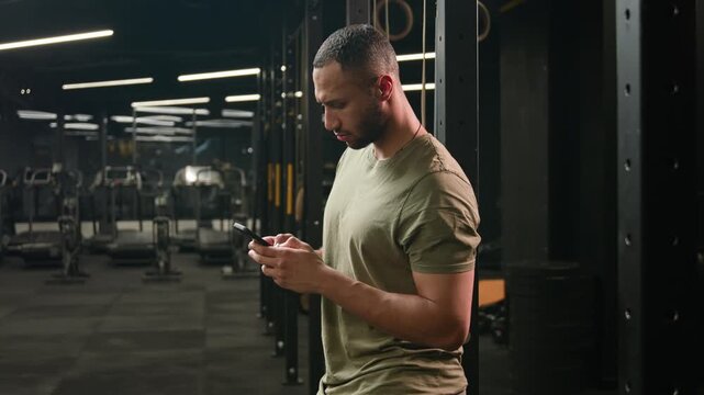 Happy Latino American male man fitness trainer using smartphone in gym looking at camera smiling athlete bodybuilder browsing mobile phone during workout break and checking social media online indoor