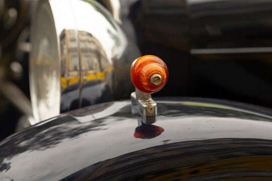 Close-up of an amber glass hood ornament mounted on the glossy black fender of a vintage car. The small round ornament sits on a chrome base with reflected