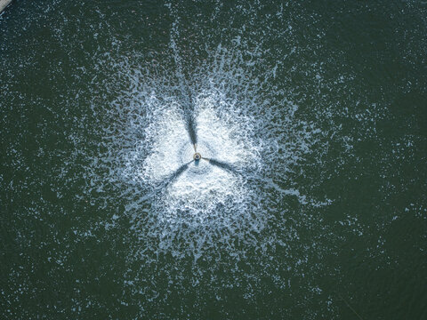 Aerial view of a water aerator creating a star-shaped pattern of bubbles and ripples on the surface of a dark pond in Poland.