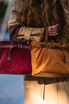 Close view of beige leather tote in woman hand with long wavy brown hair, ringed fingers, stitched seams, buckles, and tan straps during urban daytime fashion moment
