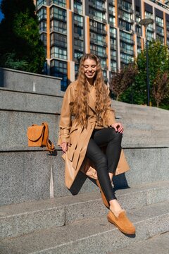 Smiling young woman seated on city steps in beige trench coat with black pants, brown boots, and tan handbag beside her in bright urban outdoor setting, with woman, smiling, female, person detail