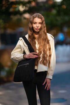 Young woman with long wavy brown hair wearing beige jacket and black tote bag outdoors in urban portrait with soft blurred background and gentle sunlight, with woman, female, young, adult detail