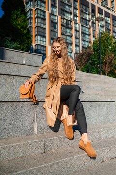 Young woman in tan trench coat sits on city steps beside orange handbag, framed by modern glass and brick architecture in daylight with relaxed smile and casual urban fashion style