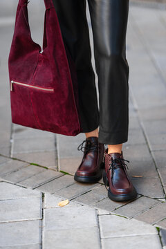 Burgundy suede handbag beside black leather pants and lace up boots on city sidewalk, creating modern urban fashion street style with rich textures and confident color contrast