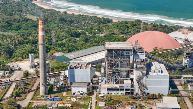 Aerial view of a power plant featuring a tall chimney, industrial structures, and a large red dome, situated near a lush coastline in Indonesia.
