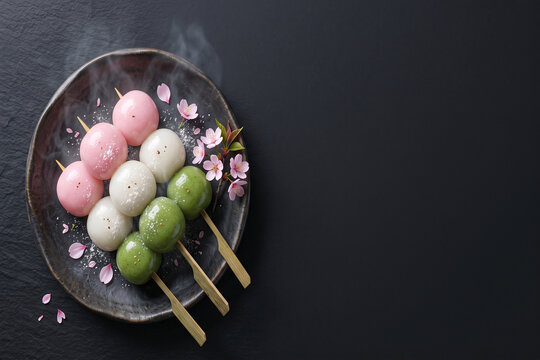 Sanshoku dango skewers plate with sakura blossoms, steam, dark background, and copy space