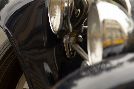 Tight view of a classic dark-painted automobile focusing on the round headlight, chrome mounting bracket, and curved fender. The glossy paint reflects