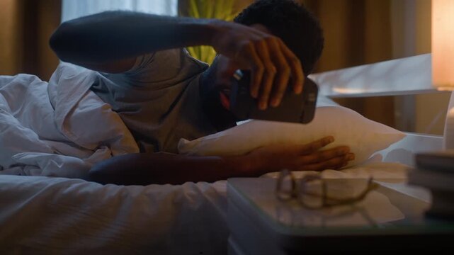 Irritated African American man waking up suffering insomnia in bed at late night tired sleepless male sleep problem exhaustion frustration digital alarm clock restlessness indoor illuminated bedroom
