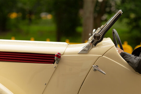 Close-up view of a cream-colored vintage car showing the passenger door, polished chrome handle, windshield frame and decorative hood vents. Soft natural