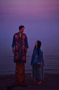 Man in traditional robe and VR visor faces woman on beach at sunset