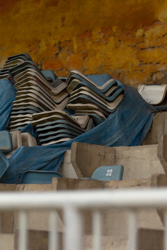 A pile of blue molded stadium seats stacked on concrete steps inside an aged, yellow-stained arena. The image shows detached seating units arranged in a heap